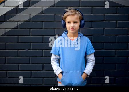 Blonde Caucasian boy in a blue hoodie and headphones stands on a city street against a gray brick wall, listening to music in a good mood. Stock Photo