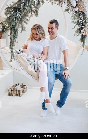 Cheerful parents with baby on xmas swing. Portrait of attractive family having fun with baby sitting on cozy swing decorated with fir branches. Stock Photo