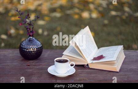 Little white cup of espresso coffee, opened book with maple leaf inside, vase with Berberis branches on rustic wooden table Stock Photo