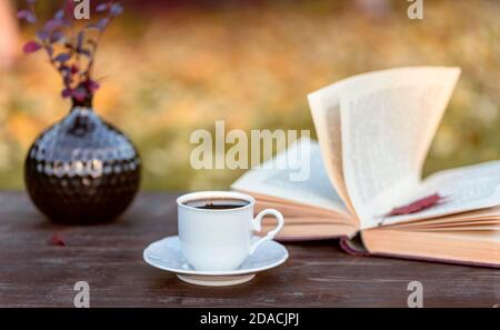 Little white cup of espresso coffee, opened book with maple leaf inside, vase with Berberis branches on rustic wooden table Stock Photo