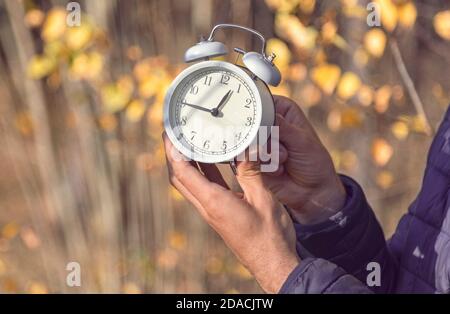 Person (hands only) adjusting time on white alarm clock in the forest on sunny autumn day. Daylight savings time concept Stock Photo