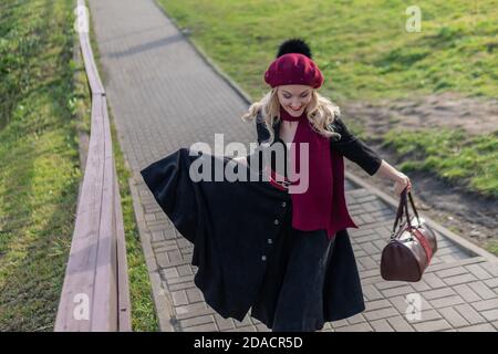 a woman walks up the stairs and in burgundy palette and biret, looking at the camera in black clothes, in the fall against a background of blue clouds Stock Photo