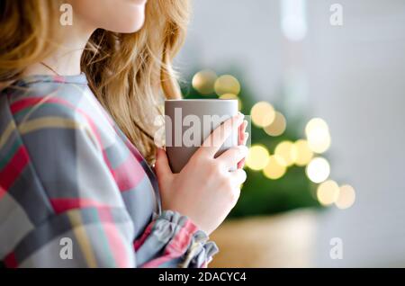 A beautiful girl in a checkered dress holds a gray cup with a hot drink in her hands. Blurred bokeh background of green Christmas tree bright lights. Stock Photo