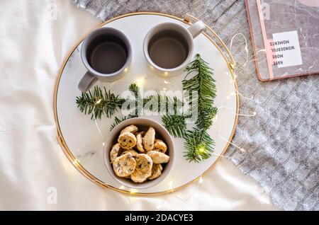 On a marble tray with a gold rim, there are two gray cups of tea and a chocolate gingerbread cookie. A treat for Santa Claus. Christmas tree branch Stock Photo