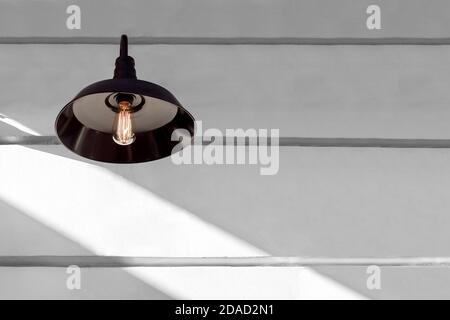 iron black loft style light with an edison lamp with threads shining warm light on a white wall with sunlight and copy space, decorative object lanter Stock Photo