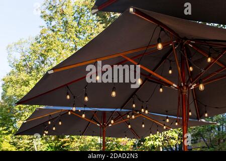 textile umbrella with a wooden frame and edison pendant lamps glowing with warm light on a backyard terrace against a background of green trees with a Stock Photo