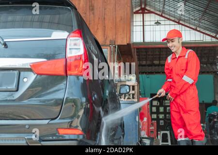 A car cleaner using a red uniform sprays high pressure water using a hose onto the tires in the car wash Stock Photo