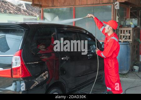 A car cleaner wearing a red uniform and a standing hat sprays water using a hose onto the roof of the car in the car wash Stock Photo