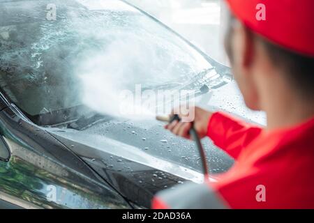 A car cleaner wearing a red uniform and a standing hat sprays water using a hose onto the windshield in the car wash Stock Photo