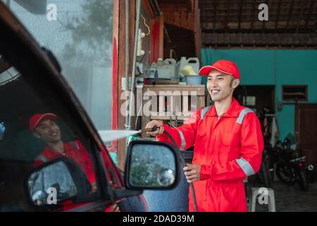 A car cleaner wearing a red uniform and a standing hat sprays water using a hose to the car in the car wash Stock Photo