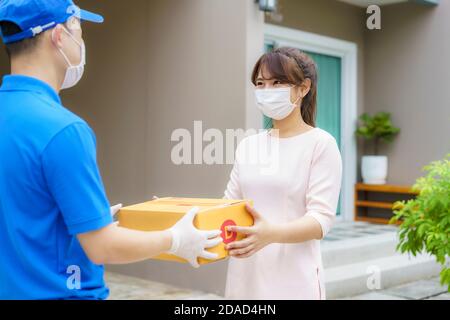 Asian delivery man wearing face mask and glove in blue uniform holding a cardboard boxes in front house and woman accepting a delivery of boxes from d Stock Photo