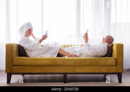 Couple in bathrobes is relaxing on the yellow sofa with mobile phones. Communication concept. Stock Photo