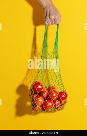 Male hand holding a green mesh bag with vegetables on yellow background with copy space. Zero waste shopping concept. string shopping bag with tomatoe Stock Photo
