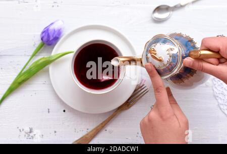 Woman pours hot red tea in a cup on a white wooden table/ Fresh blue tulip and cup of red herbal tea served on white wooden table Stock Photo