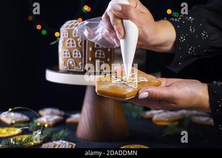 Hands of a pastry chef with a confectionery syringe for cream. Gingerbread houses, biscuits, firs, dried oranges, lights. Holiday content Stock Photo