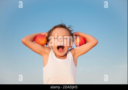 funny kid in white tank top with red apples on biceps muscle on blue sky background with copy space Stock Photo