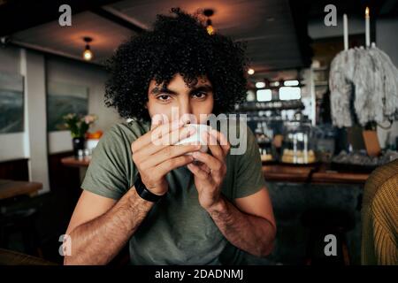 Handsome young man with curly hair sipping hot coffee relaxing in cafe Stock Photo