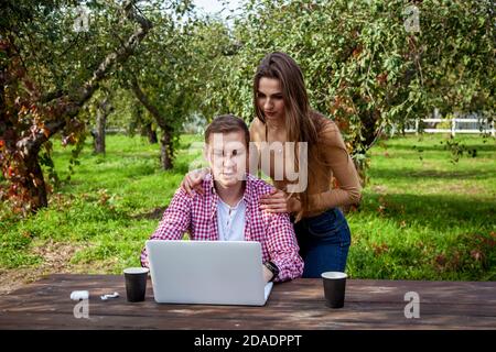 Smiling young couple spend time together in the park, study and work while sitting at a wooden table with a laptop. Guy with a girl with gadgets in th Stock Photo