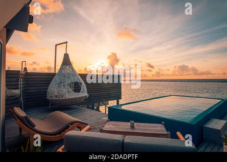Luxury resort, seascape, sunset sky and infinity pool on terrace. Sun loungers and romantic couple swing, hammock. Relaxing tropical vacation, travel Stock Photo