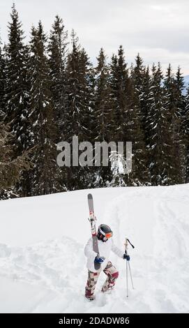 Man skier in white winter jacket holding skis and ski poles while walking the snowy path. Young man in helmet and ski goggles climbing snow-covered hill in winter mountains. Stock Photo