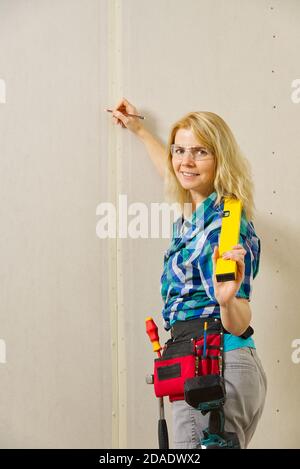 Blond woman wearing a DIY tool belt full of a variety of tools on a unpainted plasterboard wall background. Construction woman concept. Stock Photo