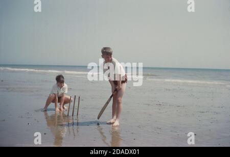 1960s, historical, two young boys in white tops and swimming trunks, playing a game of beach cricket outside on a flat, wet sandy area by the sea, England, UK. One is batting, the other is behind the make-shift stumps, wicket-keeping. In beach cricket the batter is obliged to run whenever the ball is hit regardless of how good a contact was made. A hit into the sea is generally regarded as six and out! Stock Photo