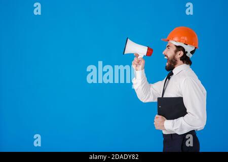 A young bearded male construction worker in a white shirt and construction helmet on a blue background. An industrial engineer-Architect shouts Stock Photo