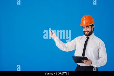 A young bearded male construction worker in a white shirt and construction helmet on a blue background. industrial engineer-Architect hold clipboard Stock Photo
