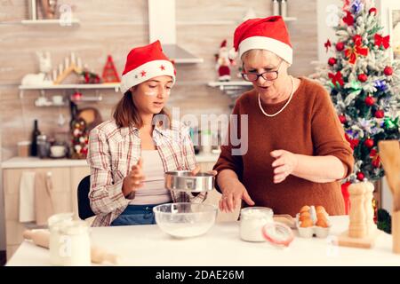 Child on christmas day with xmas tree in the background baking traditional cookies. Happy cheerful joyfull teenage girl helping senior woman preparing sweet biscuits to celebrate winter holidays wearing santa hat. Stock Photo