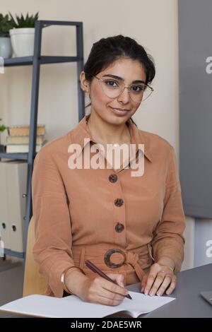 Portrait of young businesswoman smiling at camera while writing business plans in note pad sitting at the table Stock Photo