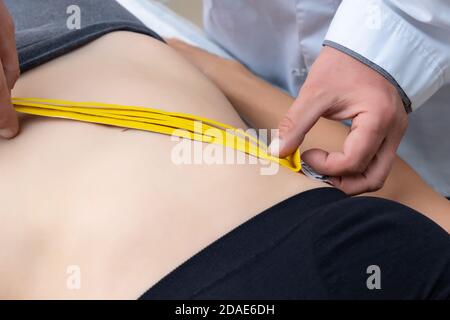 Male physiotherapist applying yellow adhesive elastic therapeutic tape on female abdominal muscle of young adult slim sporty woman lying on couch Stock Photo
