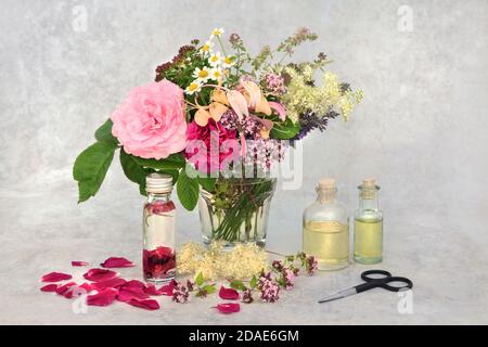 Aromatherapy essential oil preparation with summer herbs & flowers in a vase for infusing in oil. Still life with bottles and scissors on mottled grey Stock Photo