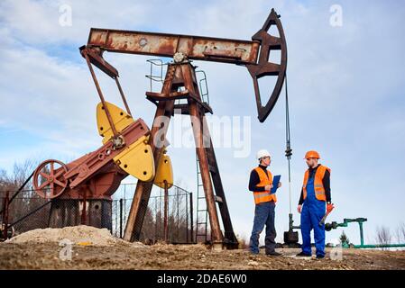 Two petroleum engineers in helmets and work vests standing on territory of oil field with oil well pump jack and sky on background. Oil man holding clipboard and talking with colleague at oil field. Stock Photo