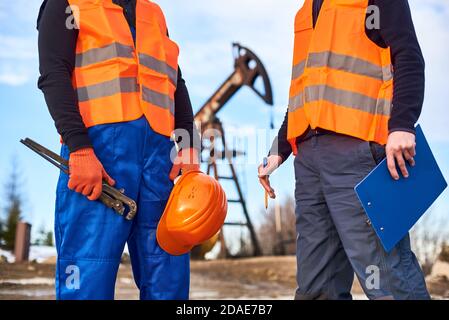 Close up of petroleum operator holding industrial wrench, helmet beside colleague with clipboard. Two oil men in working clothes standing on territory of oil field with pump jack on blurred background Stock Photo