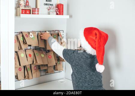 Little girl looks at the christmas advent calendar. Child in christmas hat opens a gift. Holiday concept Stock Photo