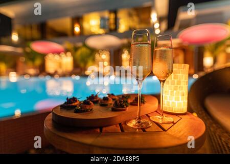 Romantic glasses of champagne on blurred luxury resort pool background of the sunset. Led lights, romantic couple appetizer, calm, night mood Stock Photo