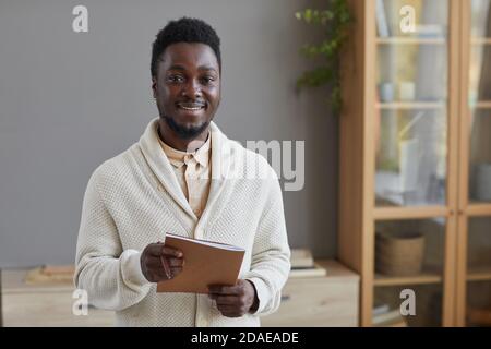 Portrait of young African businessman in white sweater holding folder and smiling at camera standing at office Stock Photo