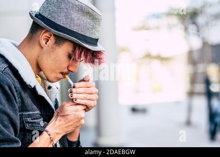 Young modern caucasian teenager male with violet hair and hat light a cigarette- smoking and smoke people concept - teen portrait in real life activity in the city - defocused background Stock Photo