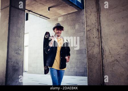 Trendy alternative young male teenager walk in the city with urban grey wall in background and skateboard - diversity concept people lifestyle with teen with violet hair and hat - portrait of colorful clothes man
