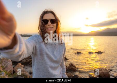 Young beautiful caucasian woman make a self portrait holding the camera at sunset on sea rocks - Smartphone selfie with water reflection of the settin Stock Photo