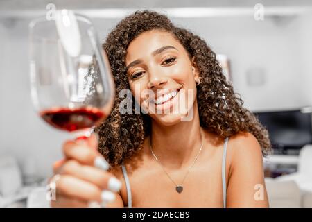Happy young Hispanic curly brunette toasting with red wine glass looking at the camera in video call conference - Beautiful black woman sitting in liv Stock Photo