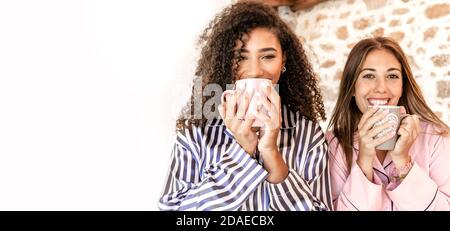 Mixed race female couple in pajama just woken up standing outdoor drinking tea looking at the camera - Two women friends smiling holding a mug with la Stock Photo
