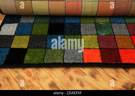 A multi-colored , rolled-up rug with a pattern of colored squares on a wooden floor. Fluffy carpet, interior element, flooring, floor covering Stock Photo