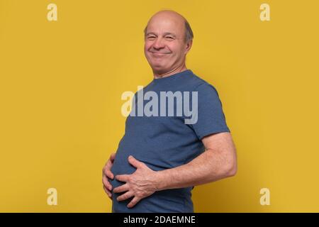 Fat senior man touching his big abdomen with hands after well eating. Stock Photo