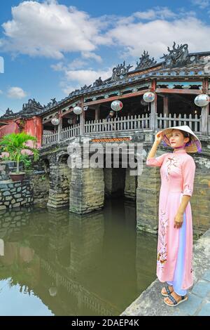 A Vietnamese young lady in traditional dress, the ao dai, posing for a photograph at the Japanese covered bridge, Hoi An, Vietnam, Asia