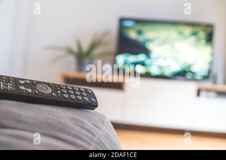 TV remote control in the foreground, tv in the blurry background. Streaming. Stock Photo