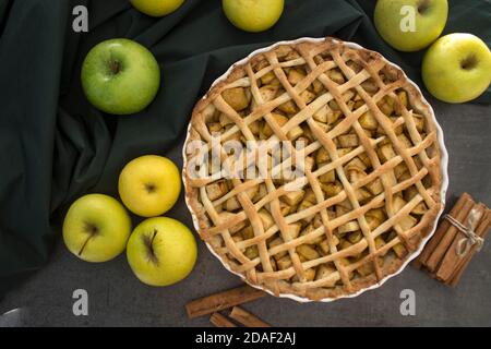 Apple pie on green fabric background. Fresh yellow apples, cinnamon sticks on a table. Autumn dessert ideas. Stock Photo