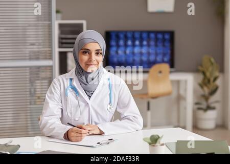 Portrait of young female doctor in white coat looking at camera while sitting at the table at office Stock Photo