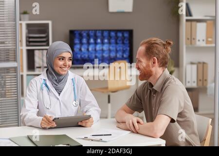 Muslim doctor using tablet pc and talking with male patient while they sitting at the table during his visit at hospital Stock Photo