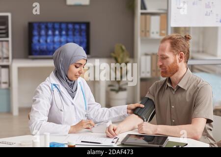Muslim female doctor measuring the pressure of male patient during his visit at hospital Stock Photo